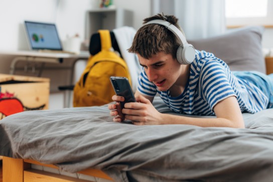 Tiener ligt op bed met koptelefoon en kijkt geconcentreerd naar zijn smartphone in een slaapkamer. (vergroot foto)