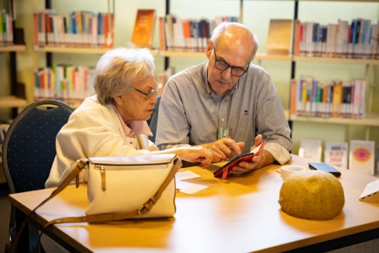 Twee oudere mensen zitten aan een tafel in een bibliotheek, verdiept in een gesprek met een tablet. (vergroot foto)